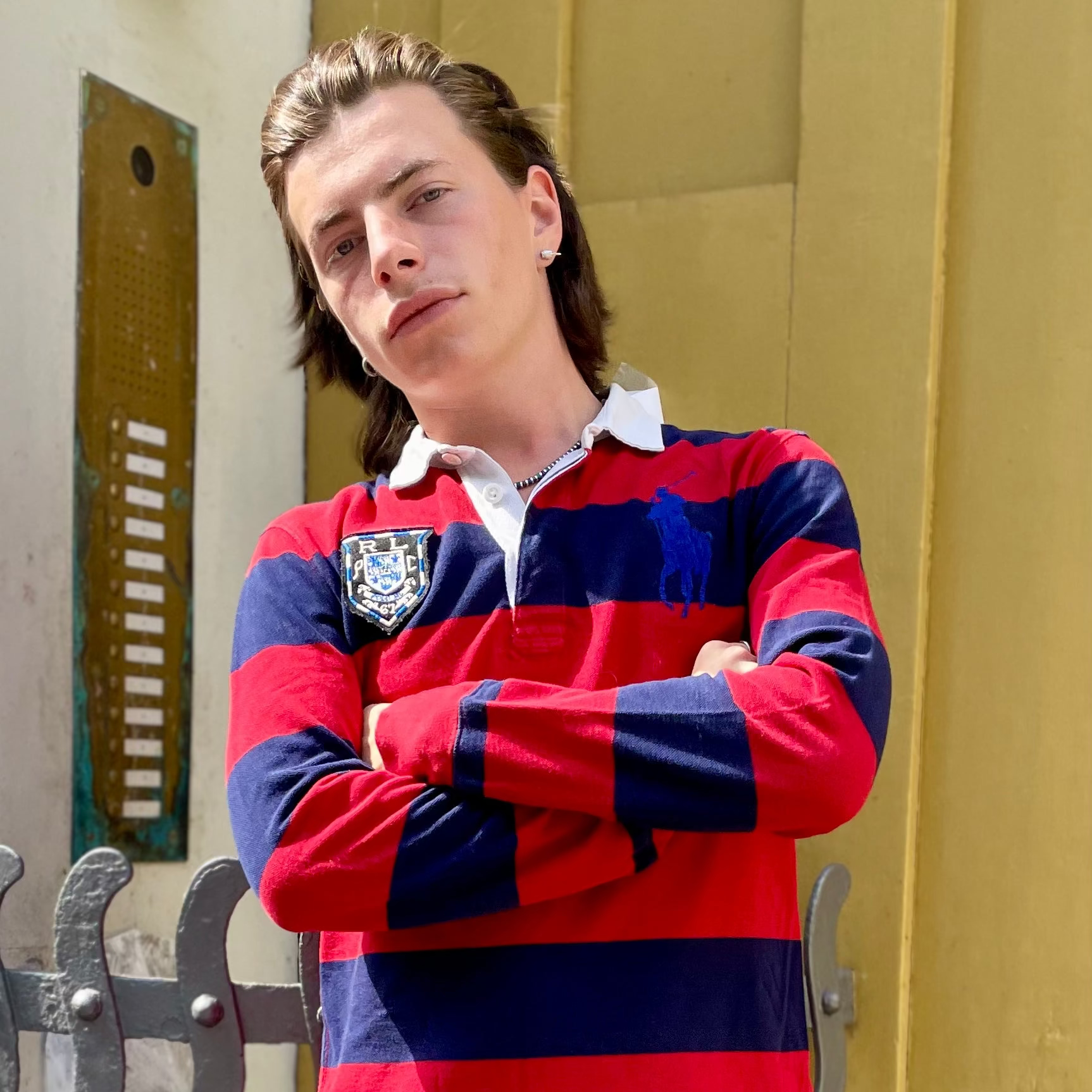 A young man with light brown hair stands with arms crossed, wearing a red and navy blue striped long-sleeve rugby shirt with a white collar, set against a yellow wall background.