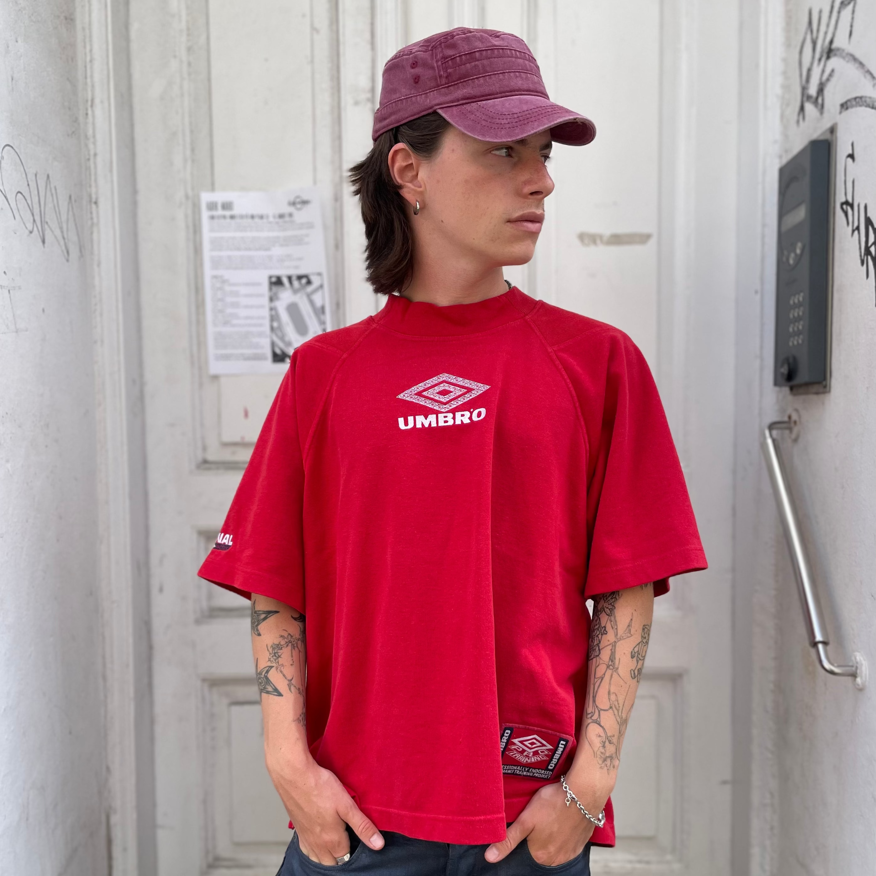 A person wearing a red Umbro t-shirt, a dusty purple cap, and dark blue jeans, standing in front of a white door with graffiti on the walls.